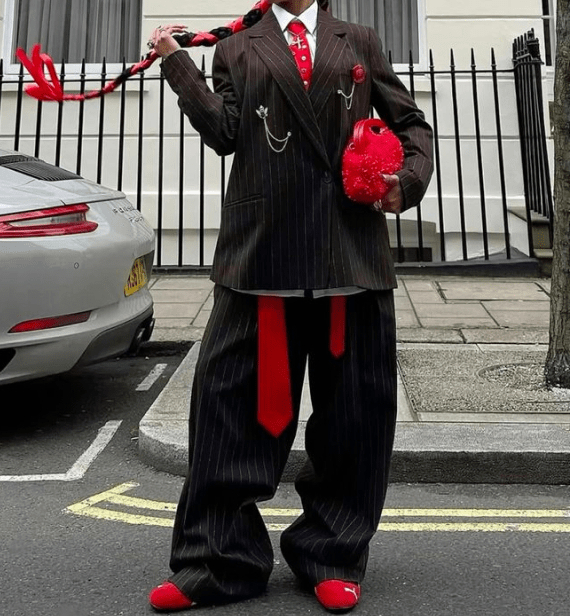A person in oversized pinstripe suiting, a red tie, and red shoes holds a textured red bag and a red-accented braid outdoors.