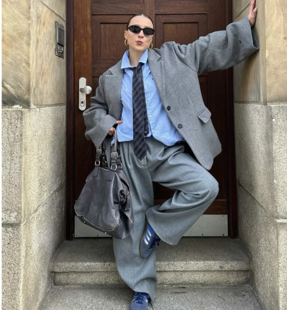 A person in oversized gray suiting, a light blue shirt, a loosened striped tie, and sunglasses poses in a doorway, holding a large black bag.