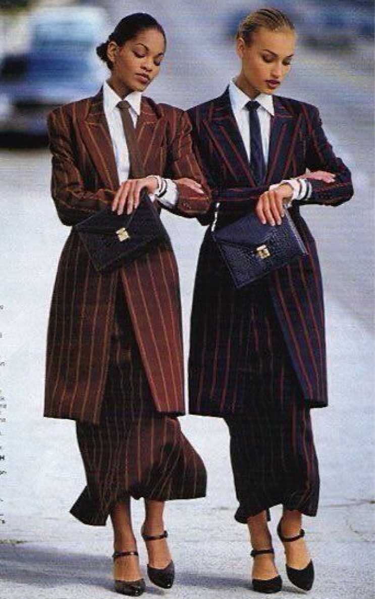 Two models in pinstriped skirt suits, one in brown and the other in navy with red stripes, pose arm-in-arm outdoors.