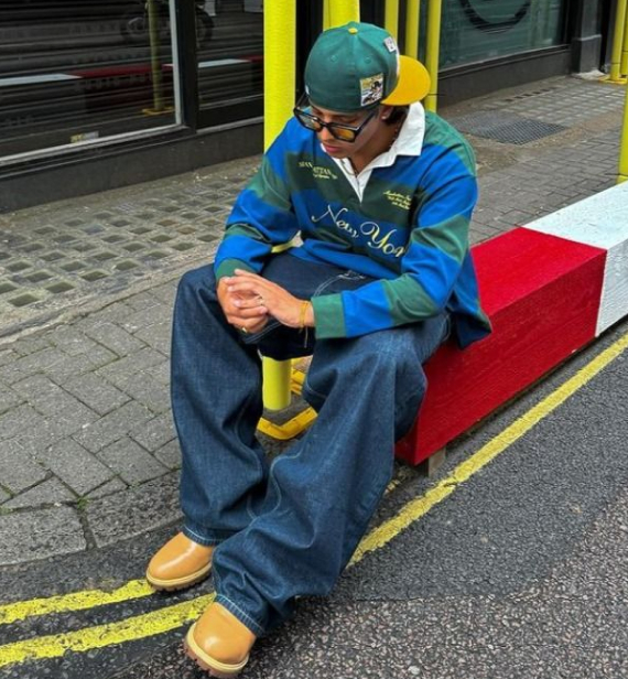 A person wearing sunglasses, a green and yellow baseball cap, a striped long-sleeved polo shirt, wide-leg jeans, and tan shoes.