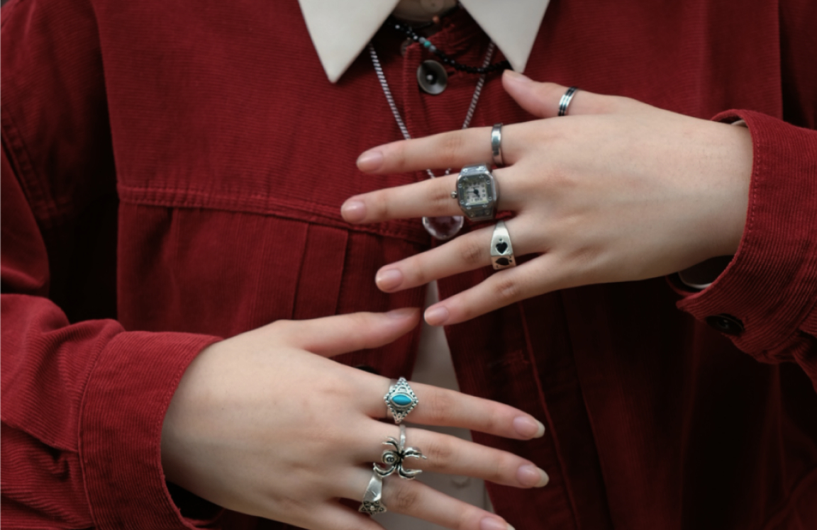 Close-up of hands wearing multiple silver rings and a digital watch stylized ring, styled with a red corduroy jacket.