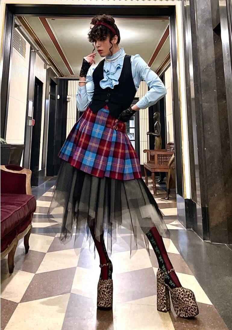 A person poses indoors wearing a layered outfit featuring a blue ruffled shirt, black vest, plaid kilt over a black tulle skirt, patterned tights, and leopard print platform shoes.
