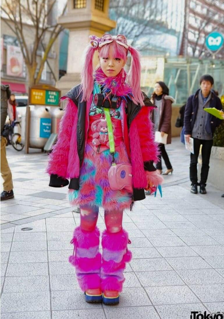 A person with bright pink hair in pigtails stands on a city sidewalk wearing a vibrant, layered Harajuku-style outfit with pink faux fur, colorful tights, and platform shoes.