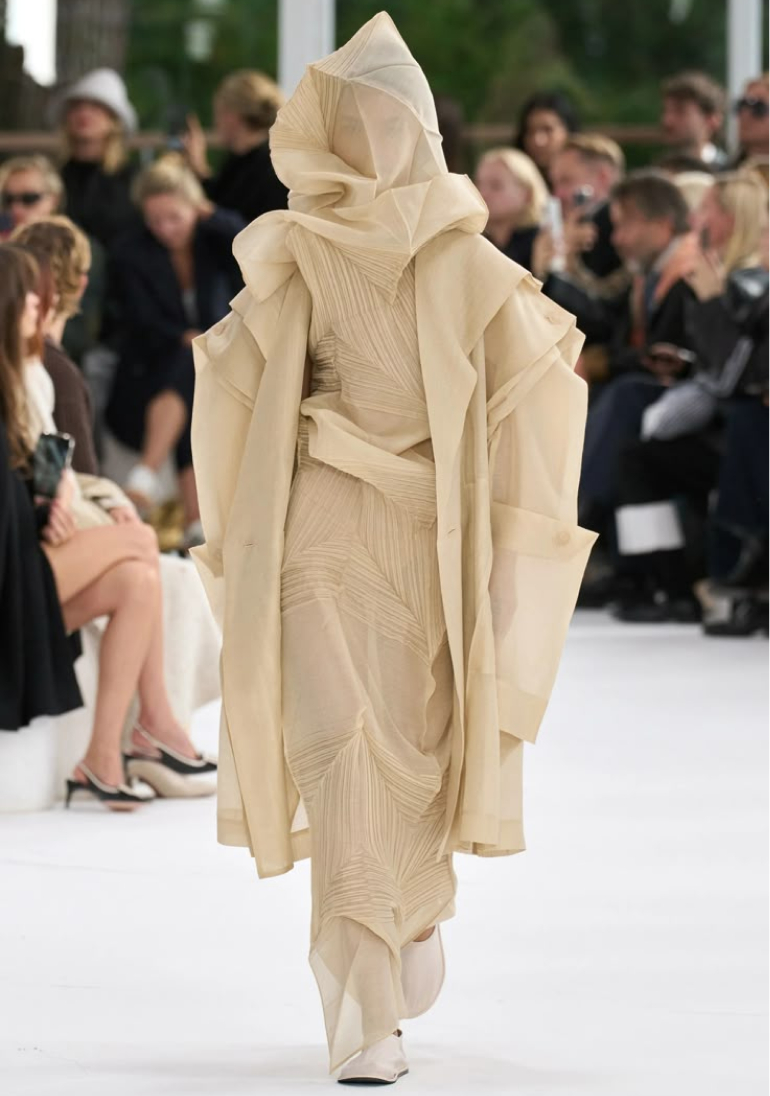 A model presents a flowing, layered beige hooded garment on a white runway before an audience.