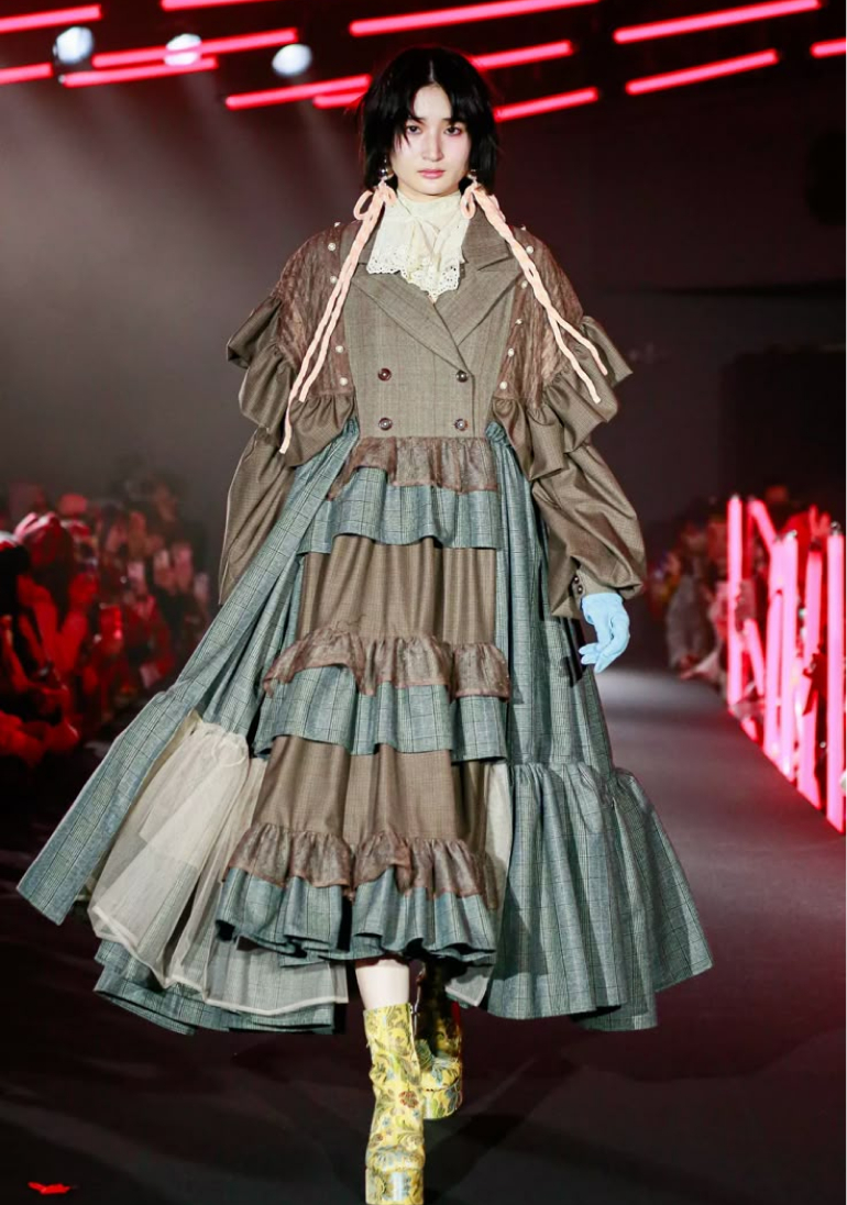 A model walks the runway in a layered, textured coat-dress with ruffles and unique detailing, paired with patterned boots and light blue gloves.
