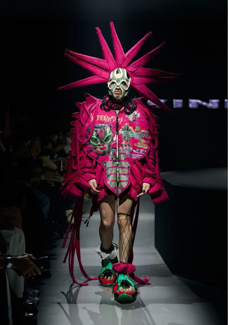 A model presents a bold, spiky pink avant-garde fashion look on a runway.