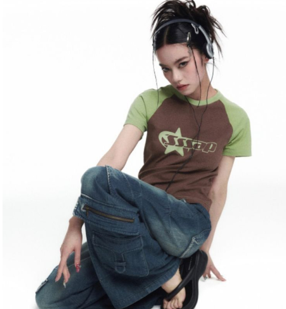 A person with dark hair and headphones poses in a brown and green graphic tee and wide-leg denim cargo pants.