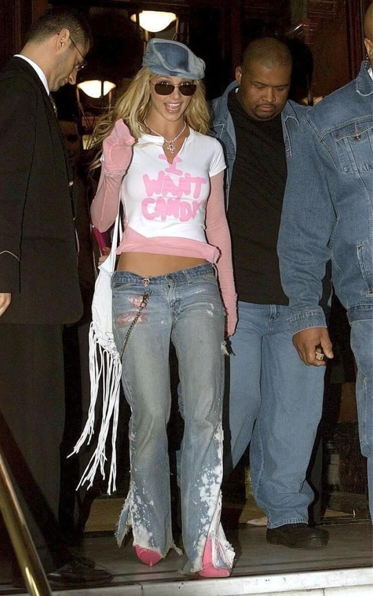 Britney Spears, wearing a 'I Want Candy' t-shirt, low-rise jeans with flared bottoms, a blue newsboy cap, and sunglasses, is surrounded by people as she walks indoors.